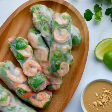 A wood serving plate containing Thai Spring Rolls with peanut sauce, limes and veggies around it