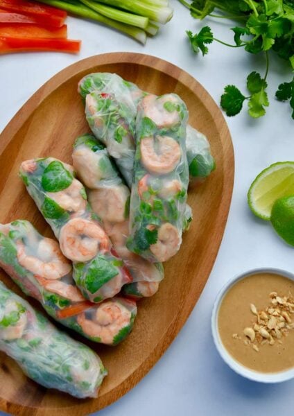 A wood serving plate containing Thai Spring Rolls with peanut sauce, limes and veggies around it