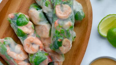 A wood serving plate containing Thai Spring Rolls with peanut sauce, limes and veggies around it