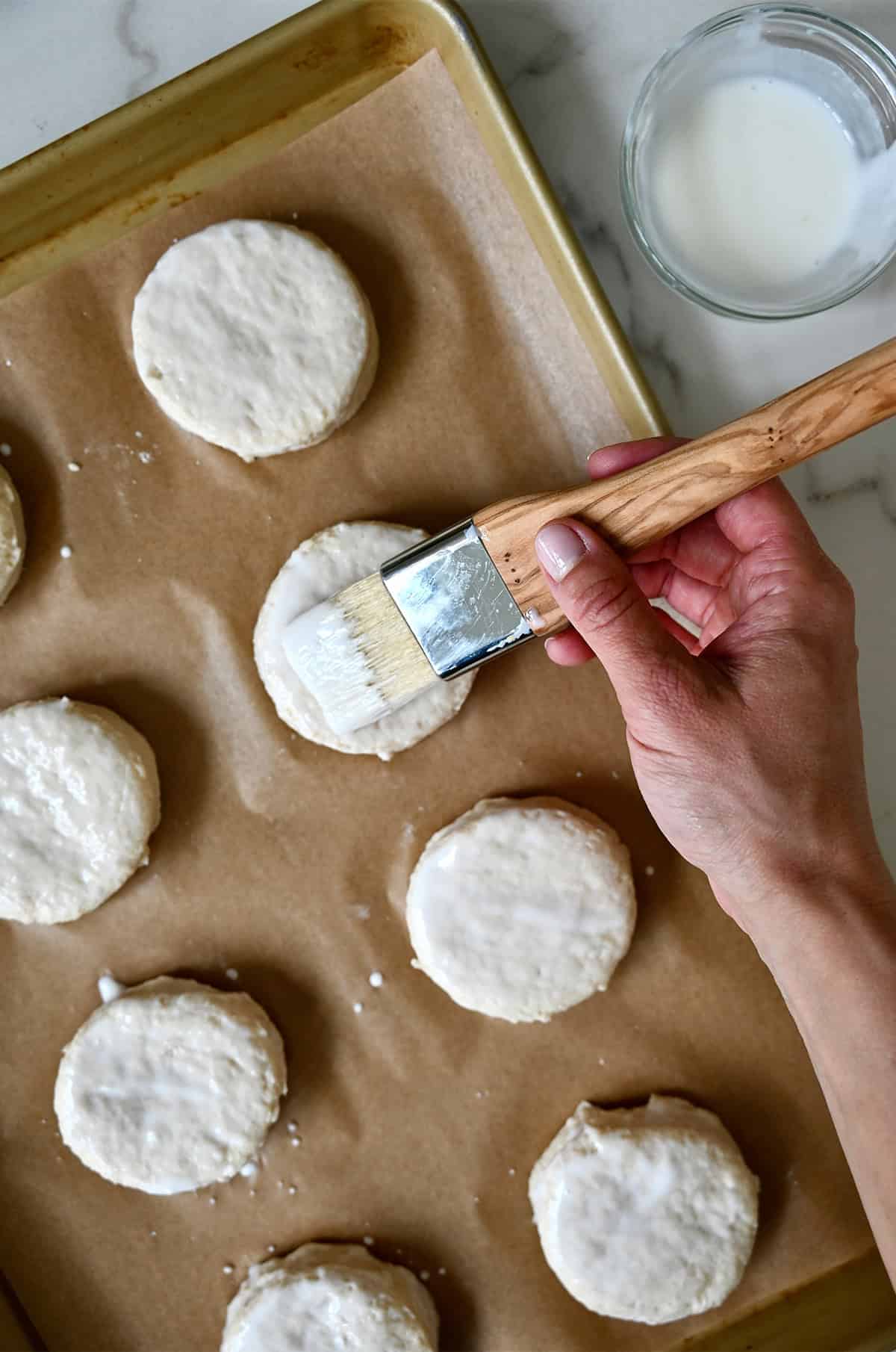Brushing buttermilk atop biscuit dough rounds on a parchment paper-lined baking sheet.