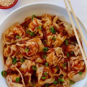 Bowl containing Spicy Chicken Wontons with chopsticks next to small red bowl with sesame seeds