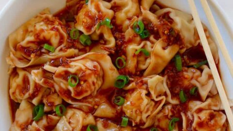 Bowl containing Spicy Chicken Wontons with chopsticks next to small red bowl with sesame seeds