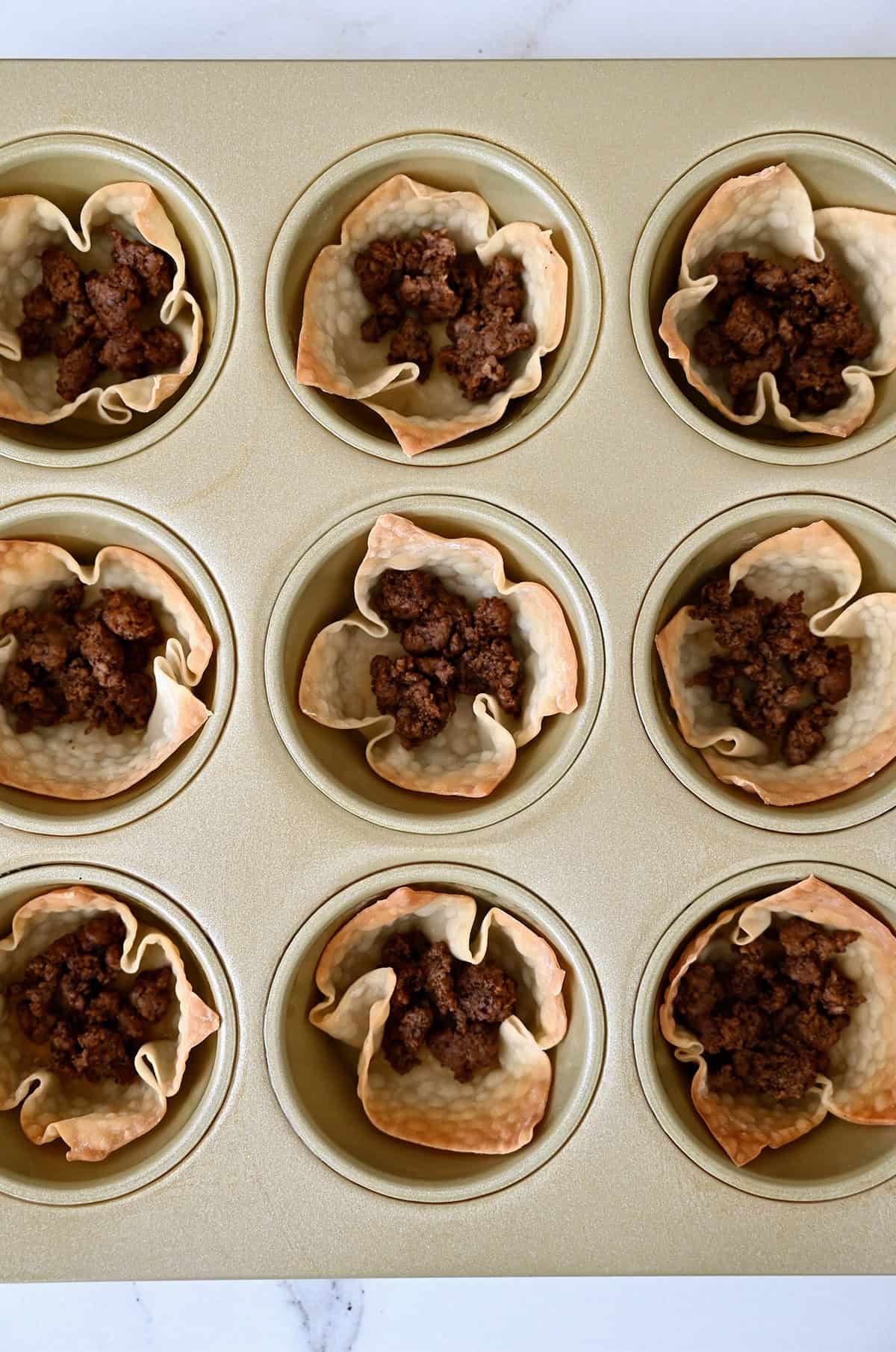 Baked wonton cups in a muffin tin, each filled with taco meat.