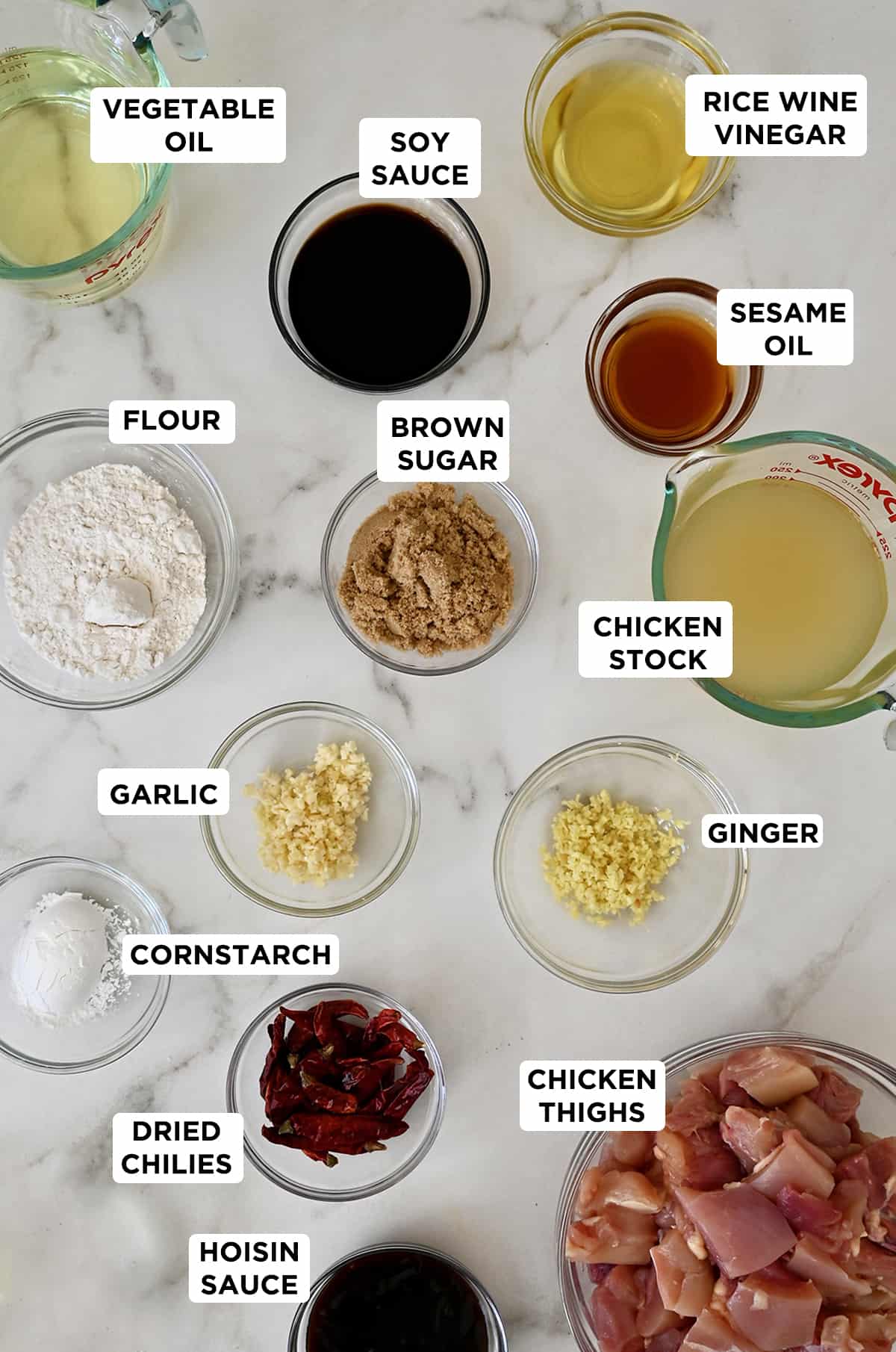 Ingredients for general Tso's chicken in various sizes of glass bowls, including vegetable oil, hoisin sauce, sesame oil, brown sugar, chicken stock, minced garlic and ginger, flour, cornstarch, chicken thigh pieces, whole dried chilies, soy sauce, and rice wine vinegar.