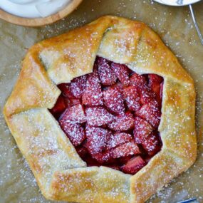 Strawberries and Cream Galette next to forks, bowl with whipped cream and sift containing powdered sugar