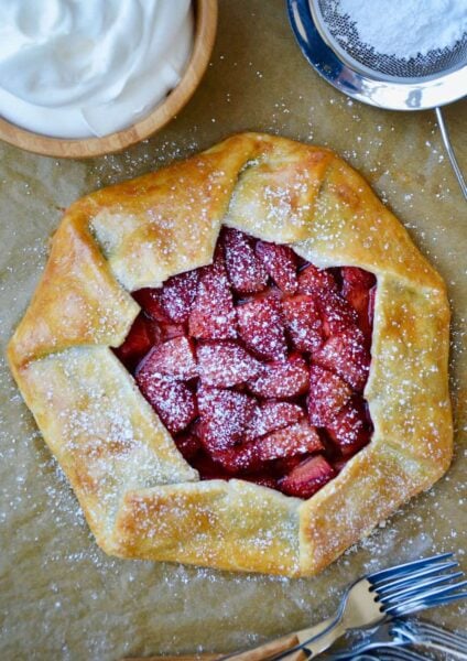 Strawberries and Cream Galette next to forks, bowl with whipped cream and sift containing powdered sugar