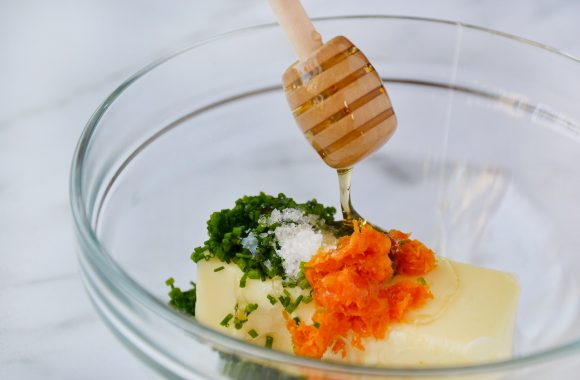 Adding honey to a glass bowl containing butter, chives, orange zest and salt