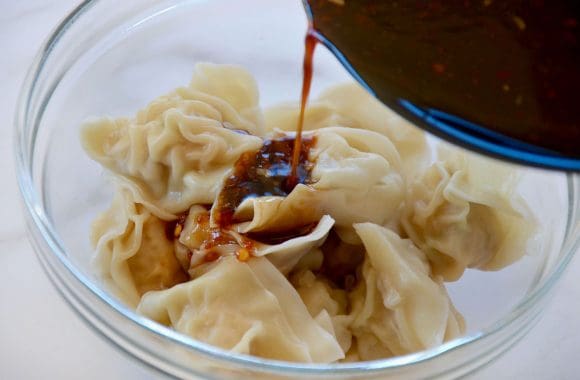 Spicy sauce being poured over wontons in glass bowl