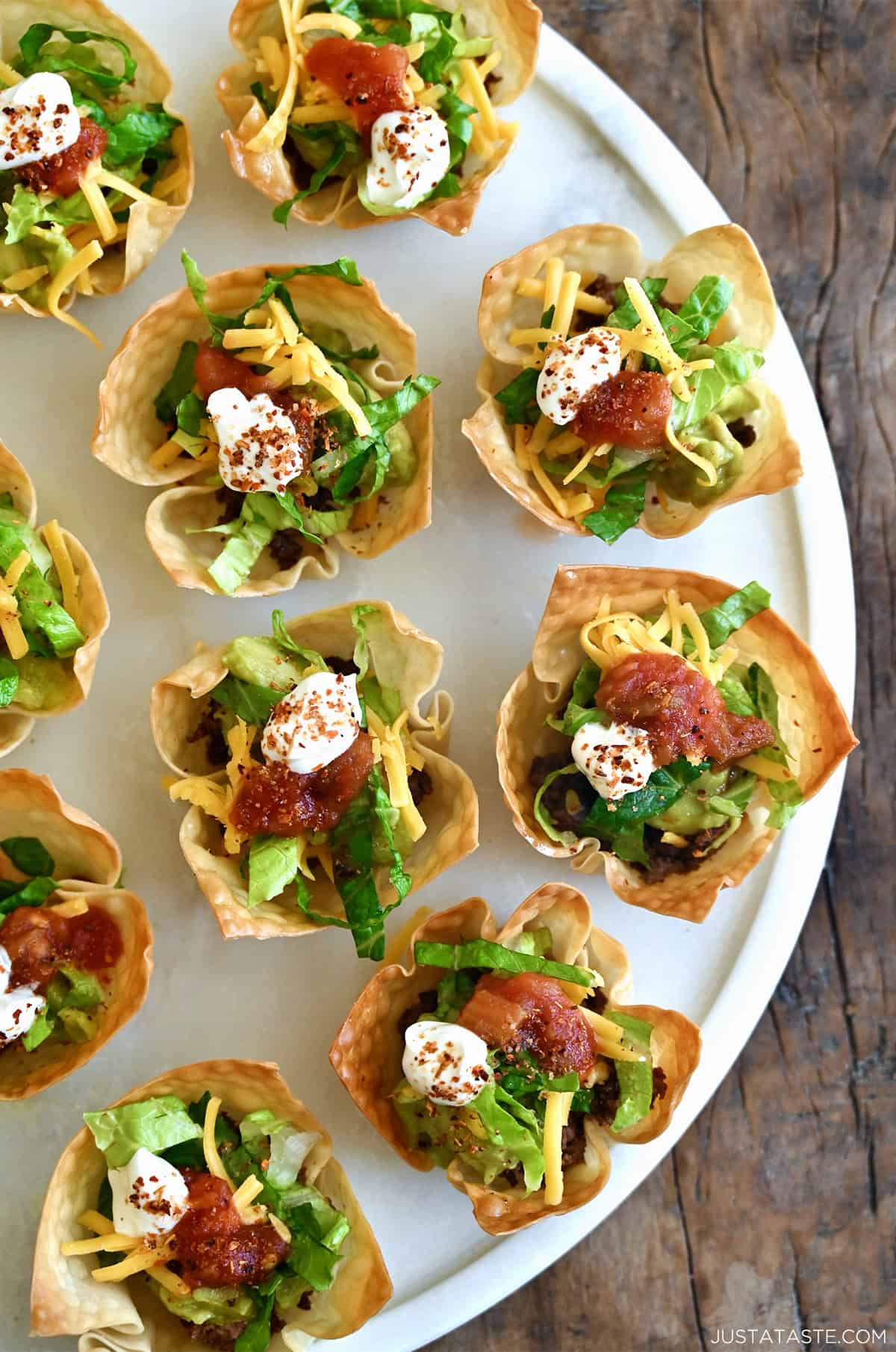 Taco salad cups topped with guacamole, shredded iceberg lettuce, salsa and a small dollop of sour cream on a serving plate.