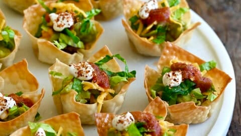 Taco cups featuring crispy baked wonton wrappers filled with seasoned taco meat and topped with shredded lettuce, shredded cheese and a dollop of sour cream.