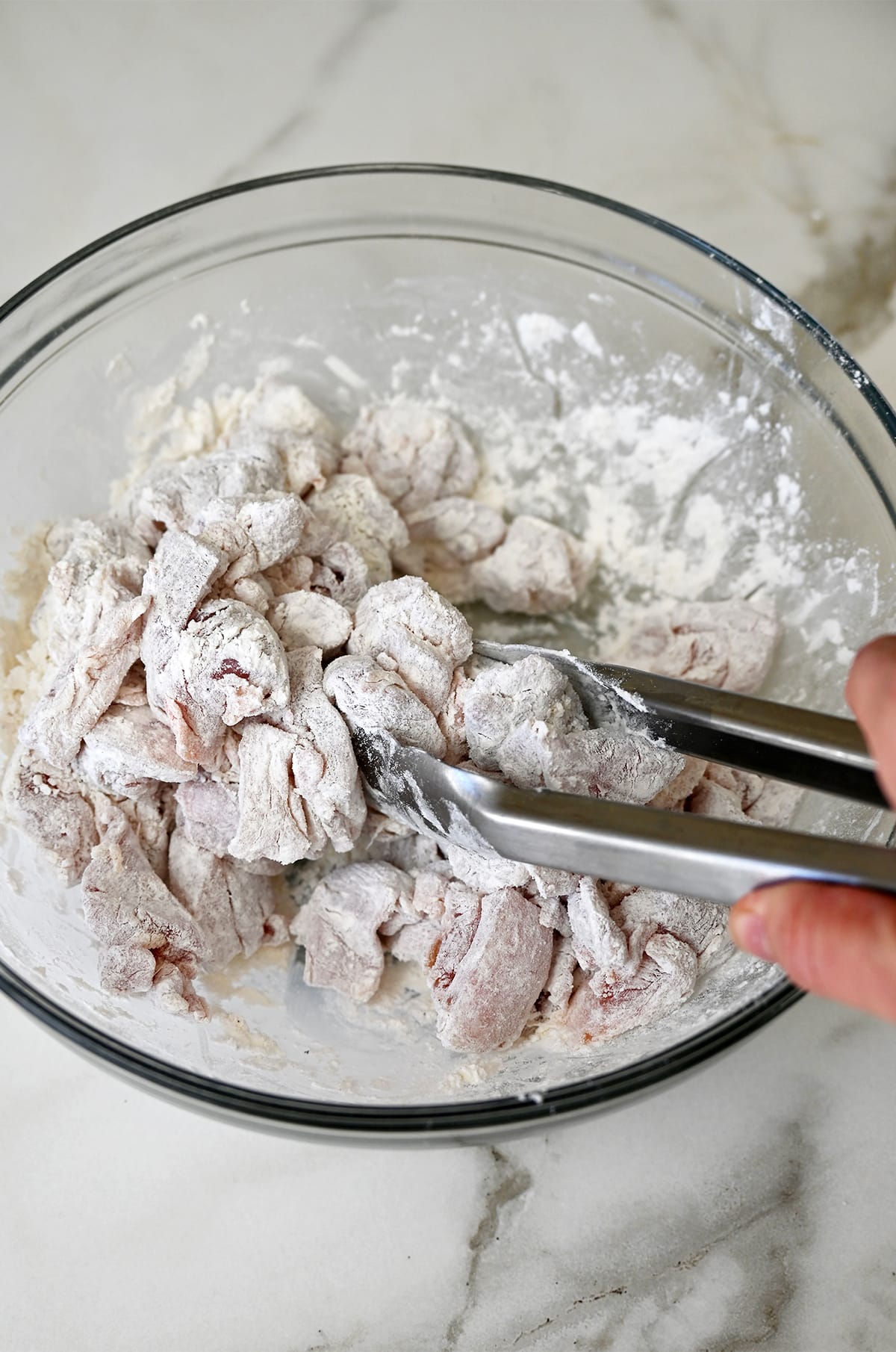 A hand holding tongs tosses chicken pieces in a mixture of flour and cornstarch in a glass bowl.