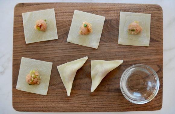 Wood cutting board with wonton wrappers, chicken mixture, and small bowl containing water 