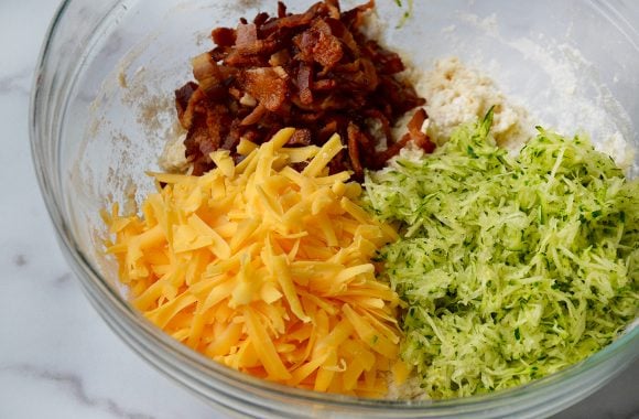 Glass bowl containing grated zucchini, shredded cheddar cheese and chopped bacon