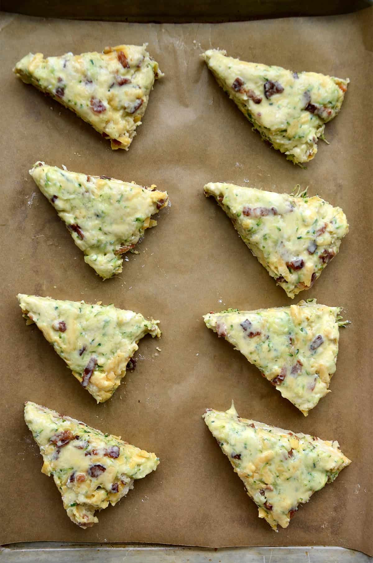 Unbaked cheddar zucchini scones brushed with buttermilk on a parchment paper-lined baking sheet.