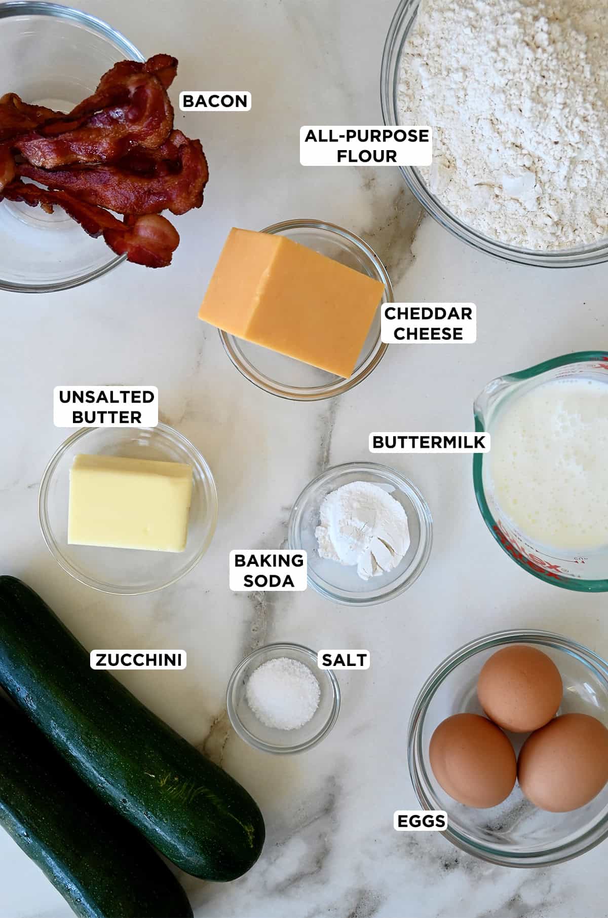 Two zucchinis next to various sizes of glass bowls containing butter, crispy bacon, cheddar cheese, flour, buttermilk, baking soda, salt and eggs.