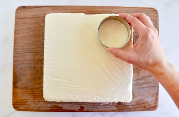 A wood cutting board containing a sheet of ice cream being cut out with a circular cookie cutter