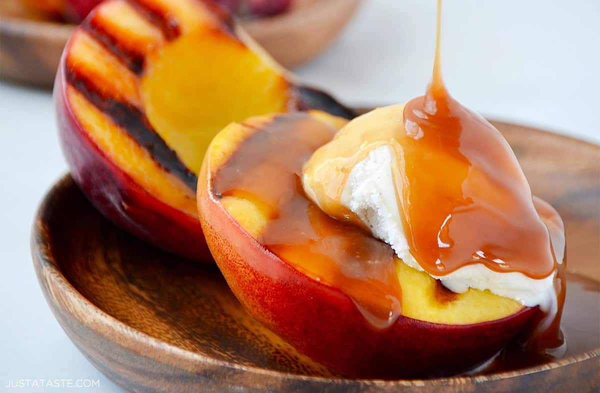 Grilled peaches with char marks topped with vanilla ice cream being drizzled with bourbon caramel sauce.