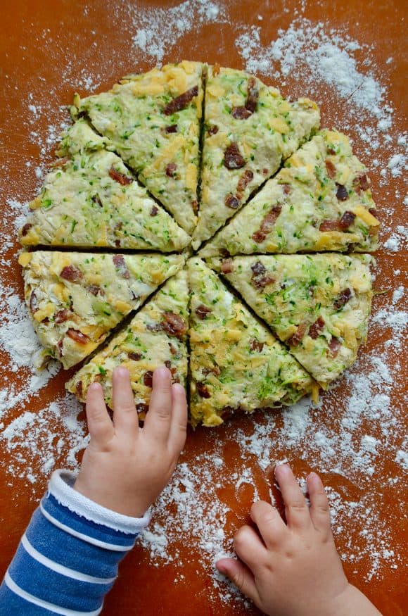 Child's hands reaching for zucchini bacon cheddar scones on generously floured cutting board