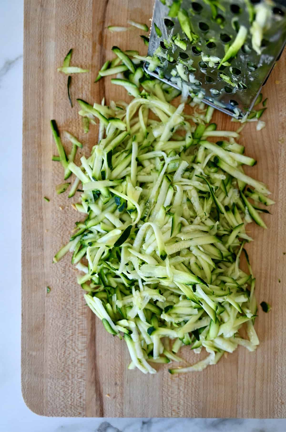 Shredded zucchini next to a box grater on a wood cutting board.