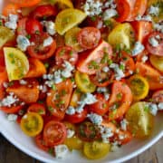 A white serving plate containing Steakhouse Tomato Salad with blue cheese crumbles, chopped fresh chives, salt and pepper drizzled with olive oil.