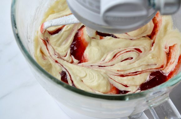 A glass stand mixer bowl containing cake batter swirled with strawberry jam