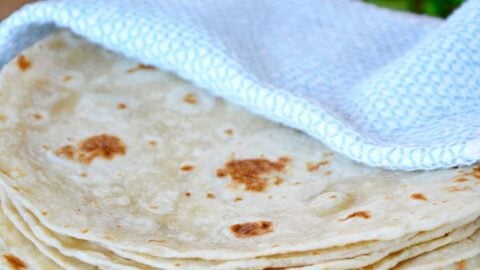 A stack of homemade tortillas in a blue towel with cheese and cilantro in the background