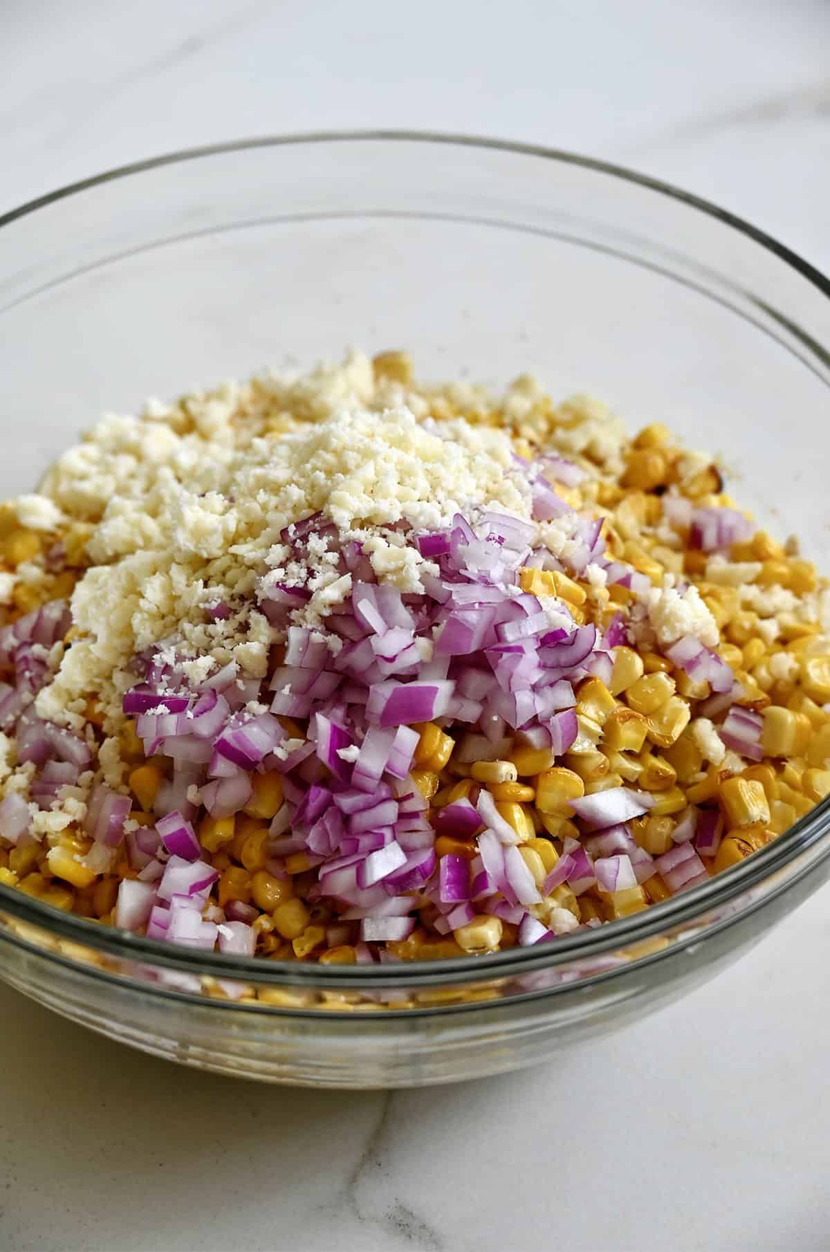 Crumbled Cotija cheese and diced red onions atop elote salad in a bowl.