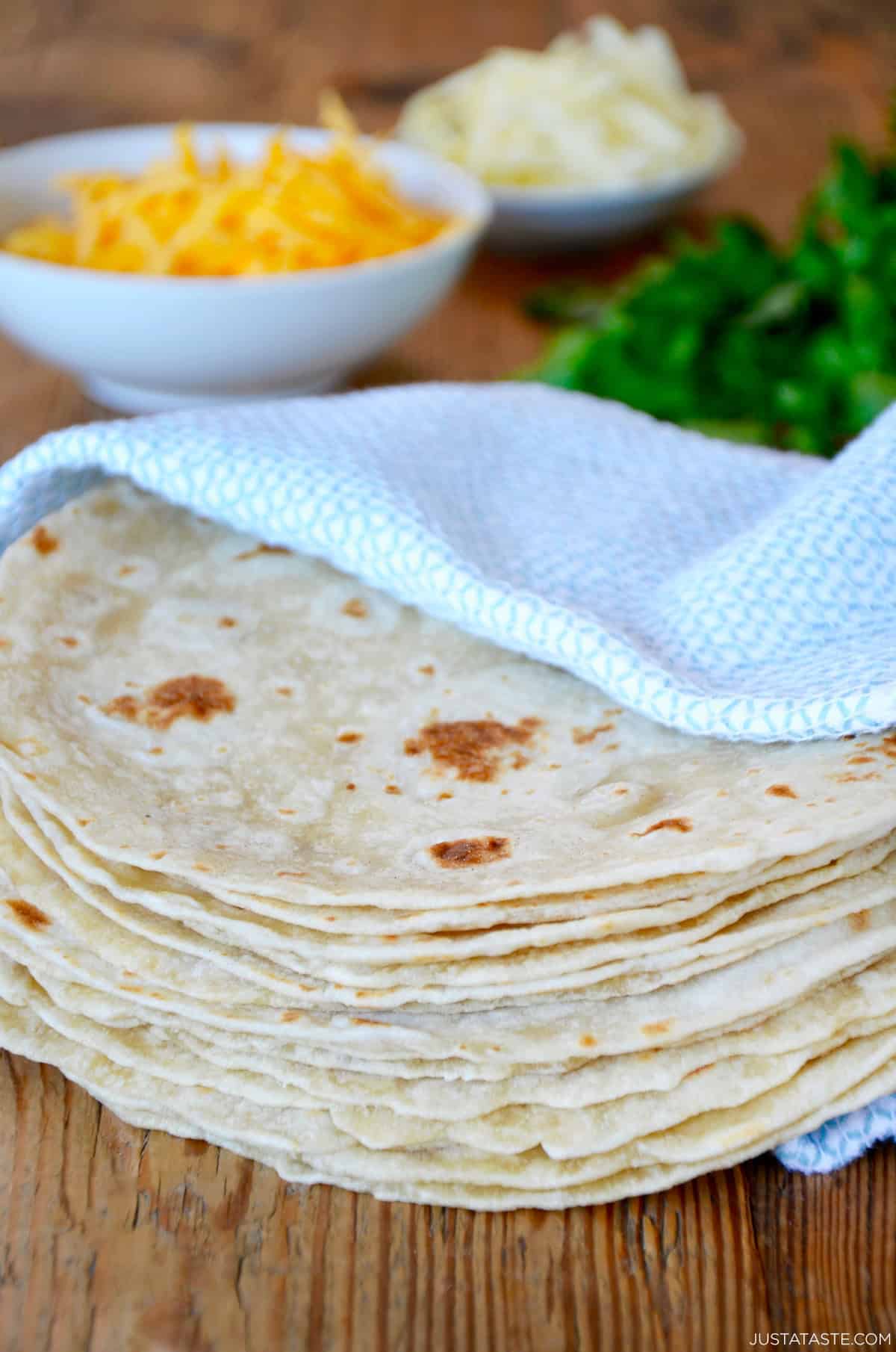 A stack of homemade tortillas in a blue towel with cheese and cilantro in the background.