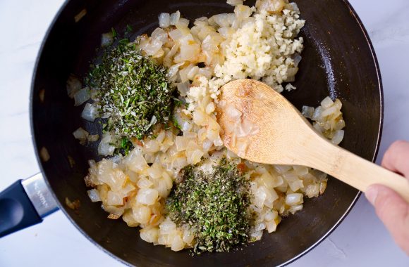 Wooden spoon stirring caramelized onions, minced garlic and fresh herbs in sauce pan