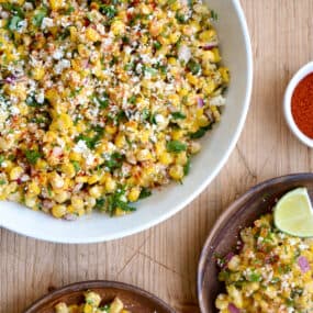 A top-down view of a large bowl containing Elote Corn Salad next to two plates loaded with the salad and a lime wedge.