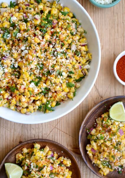 A top-down view of a large bowl containing Elote Corn Salad next to two plates loaded with the salad and a lime wedge.