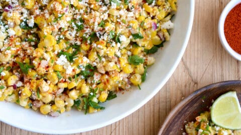 A top-down view of a large bowl containing Elote Corn Salad next to two plates loaded with the salad and a lime wedge.