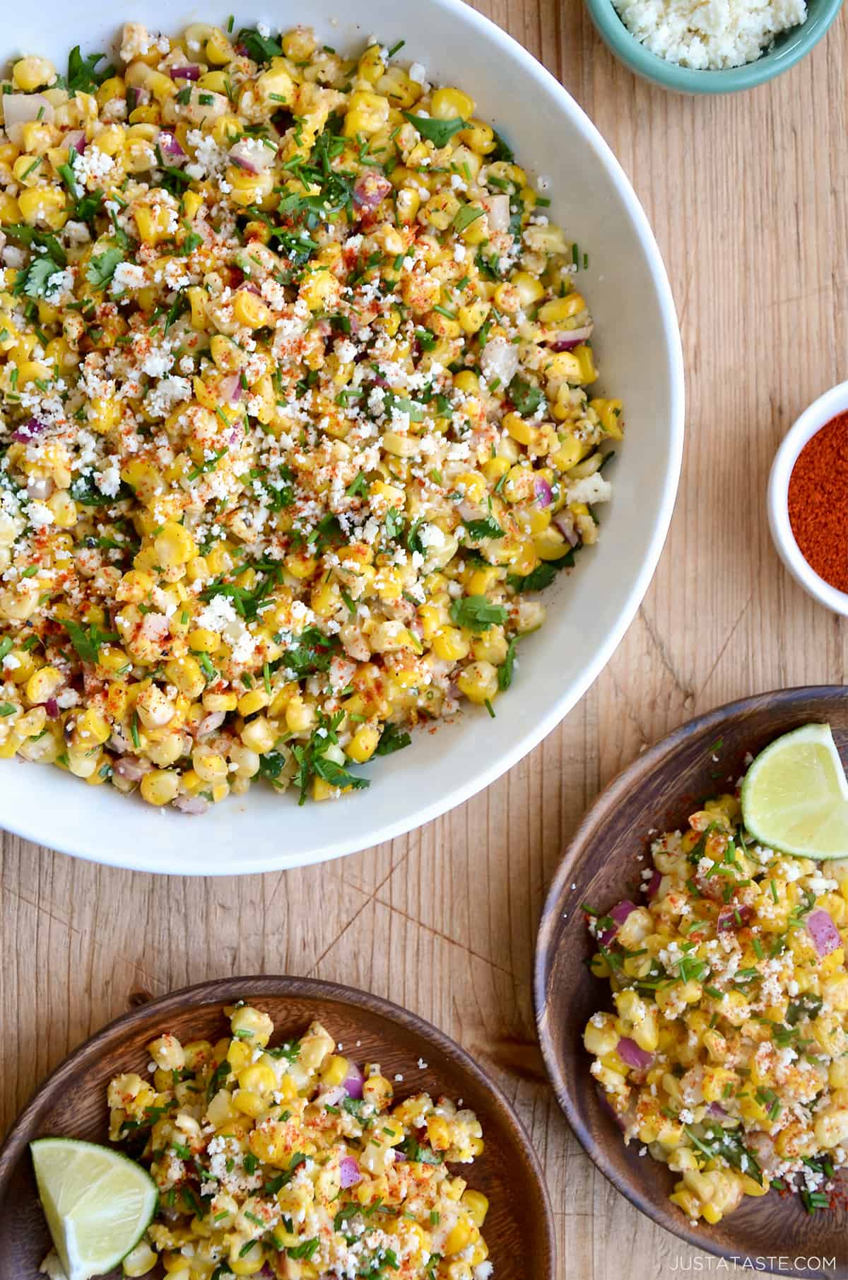 A top-down view of a large bowl containing Elote Corn Salad next to two plates loaded with the salad and a lime wedge.