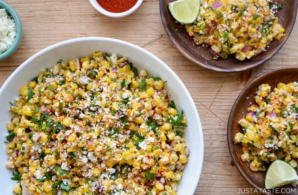 Elote in a large white bowl next to two plates with elote garnished with crumbled Cotija cheese.