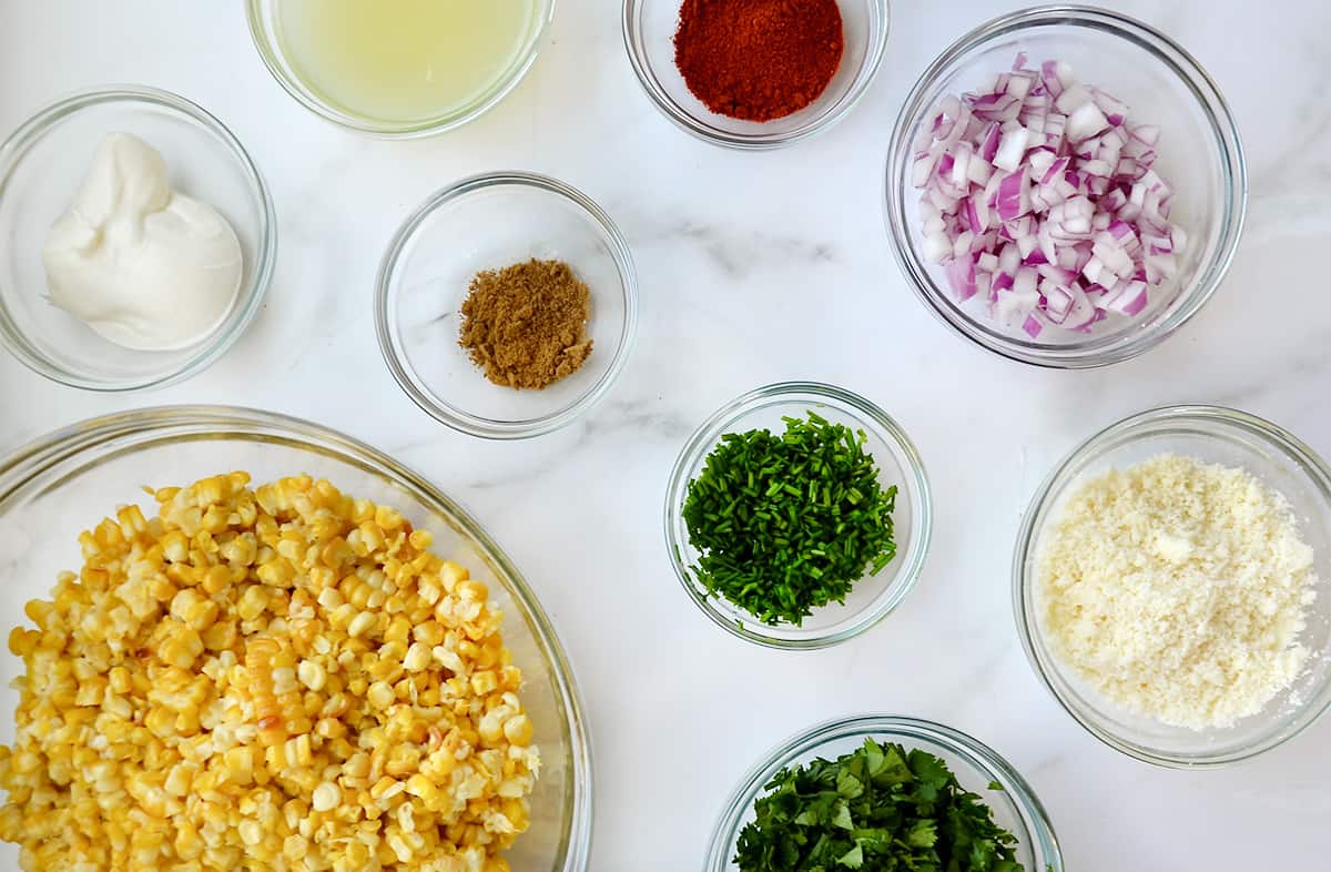 Various sizes of clear bowls containing the ingredients needed for elote salad, including corn kernels, sour cream, chives, diced red onion, cumin, paprika, cilantro and cojita cheese.