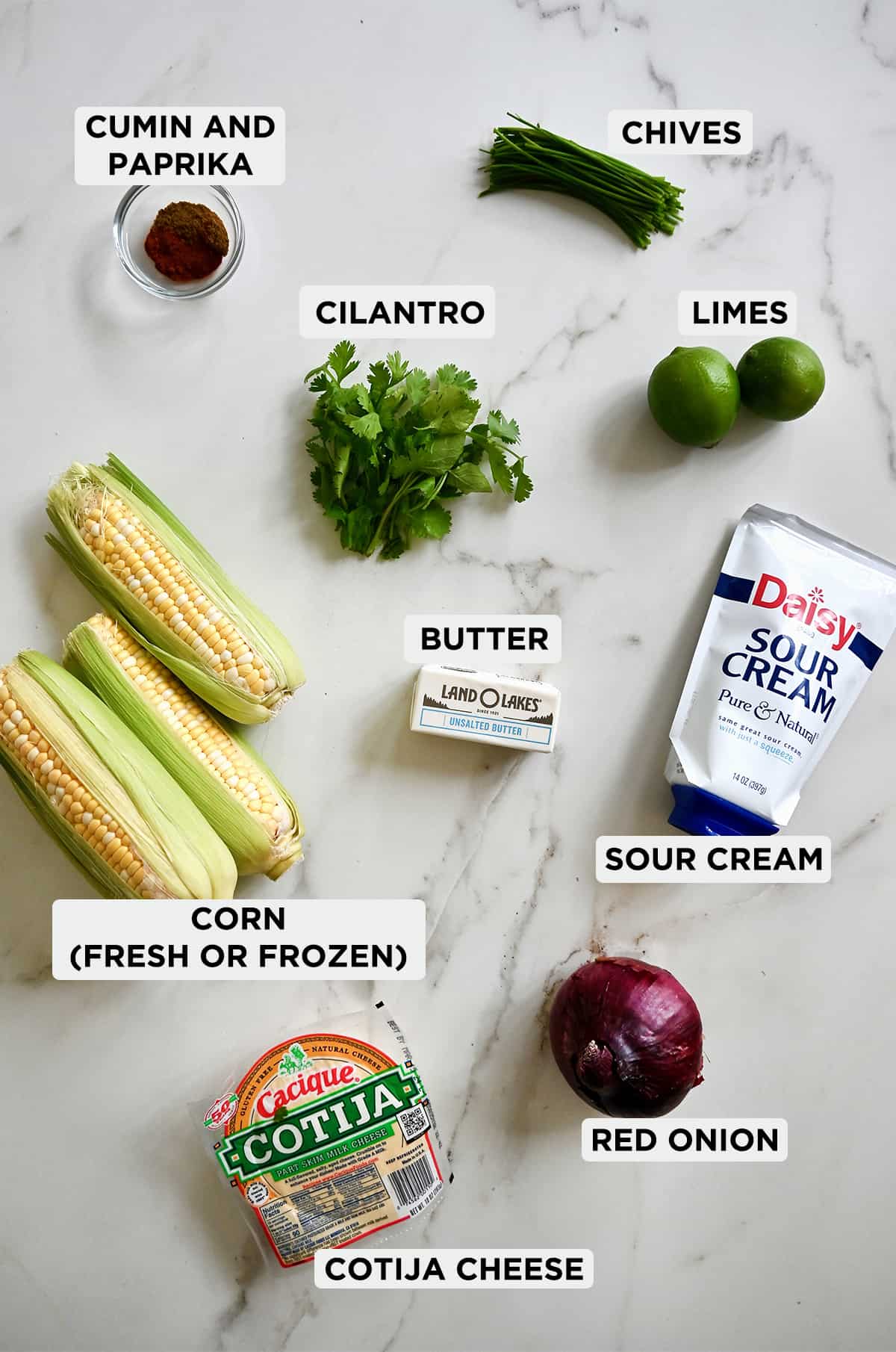 A labeled image of all the ingredients needed for Mexican street corn, including corn on the cob, cilantro, butter, cumin, paprika, red onion, limes, sour cream and cojita cheese.