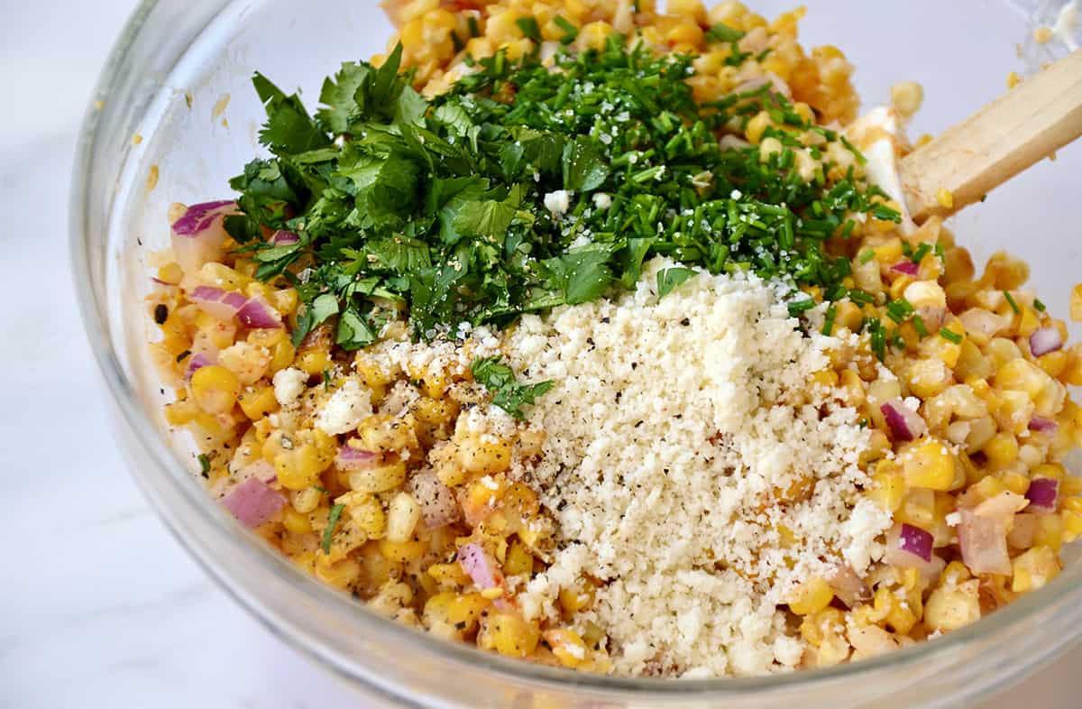 A clear bowl containing corn kernels, diced red onion, chopped cilantro and chives, and crumbled cotija cheese.