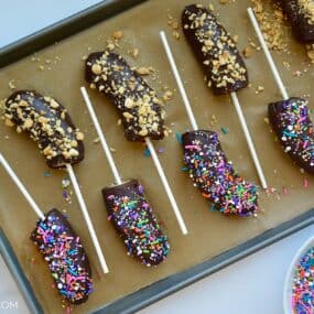 Frozen chocolate covered bananas with popsicle sticks on a baking sheet.