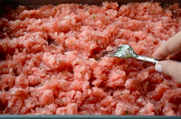 Hand holding fork scraping watermelon granita mixture on baking sheet