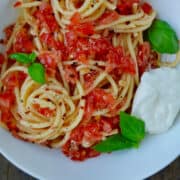 A large white bowl containing pasta with no-cook tomato sauce garnished with fresh basil and burrata cheese.