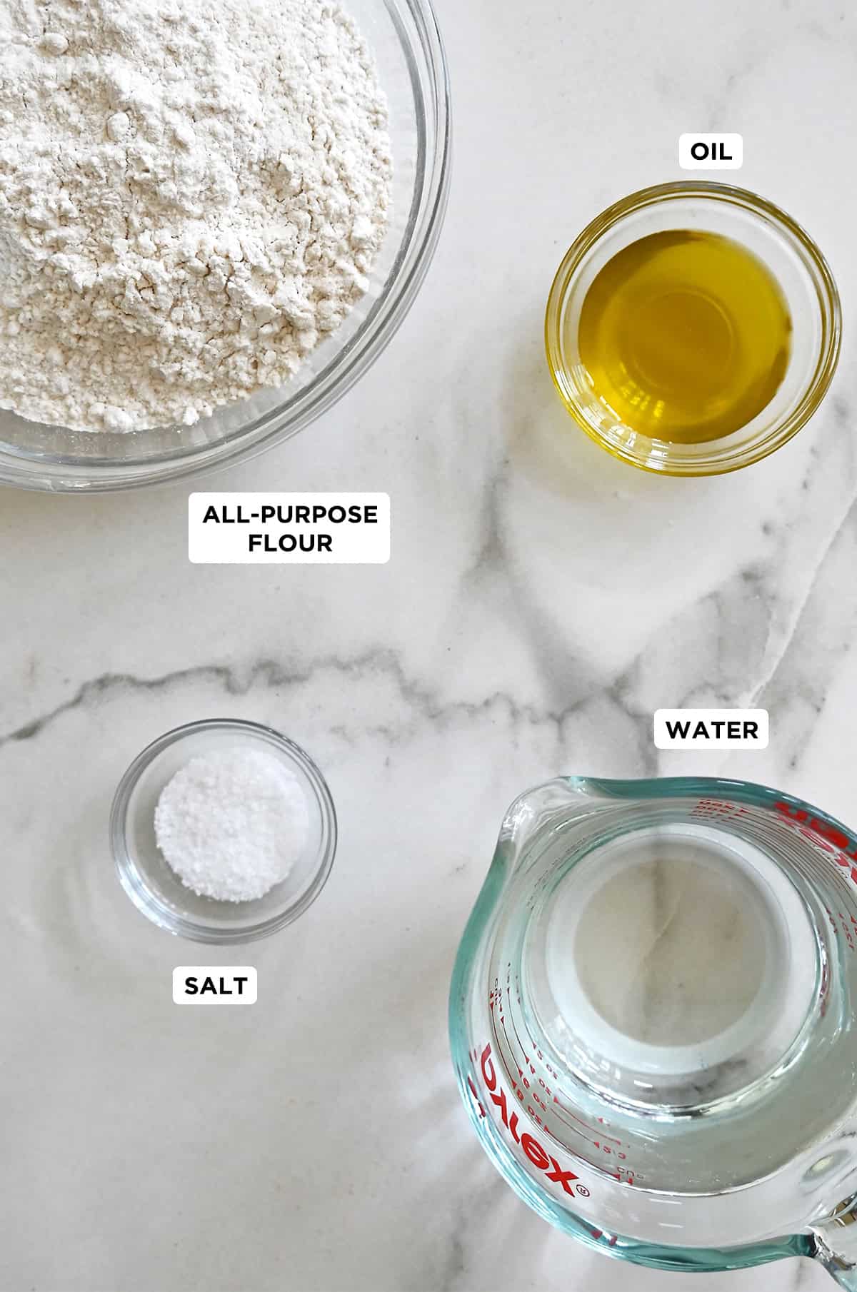 Flour tortilla ingredients, including a liquid measuring cup with water next and bowls containing salt, olive oil and flour.