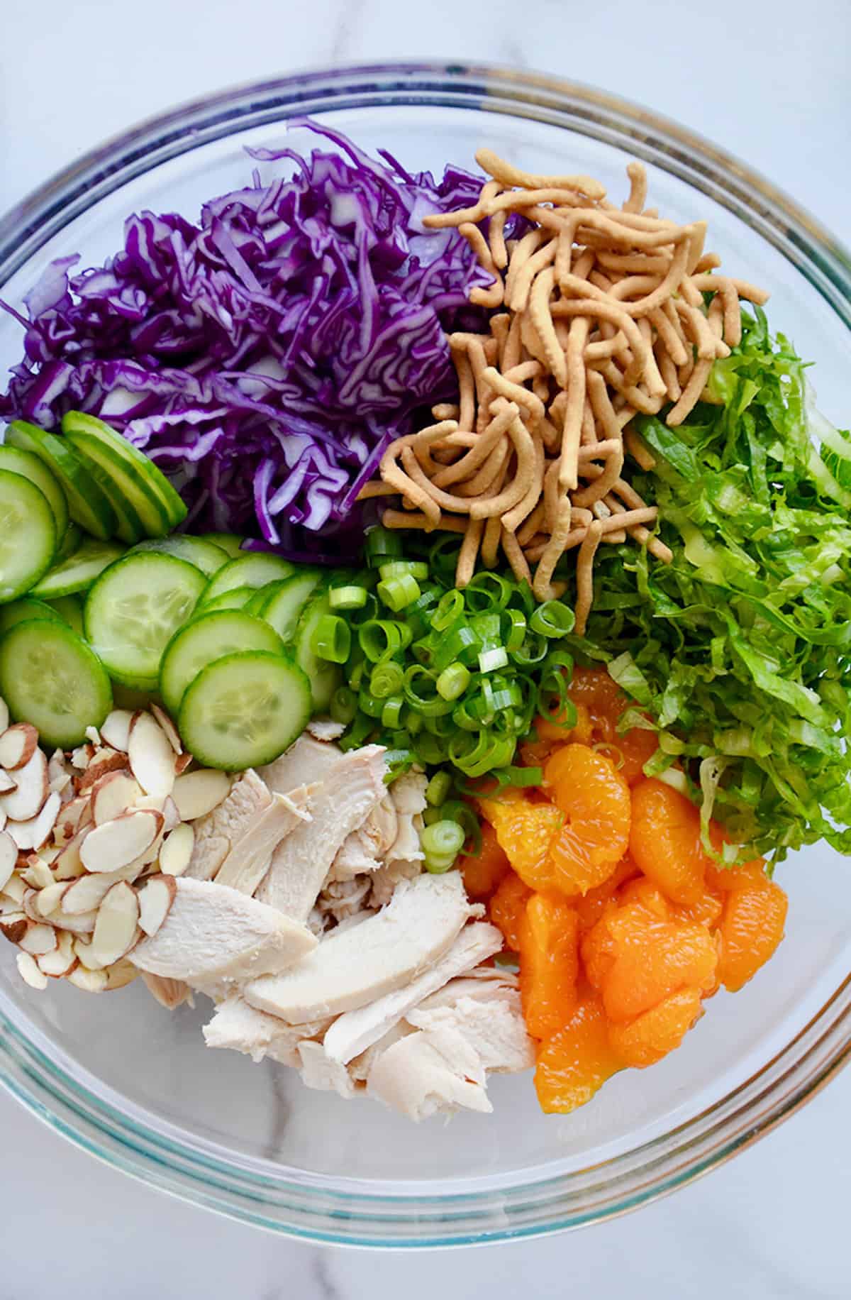 Shredded chicken breast, sliced almonds, sliced cucumbers, shredded purple cabbage, chopped scallions, crispy chow main noodles, shredded romaine lettuce and mandarin oranges in a large glass bowl.