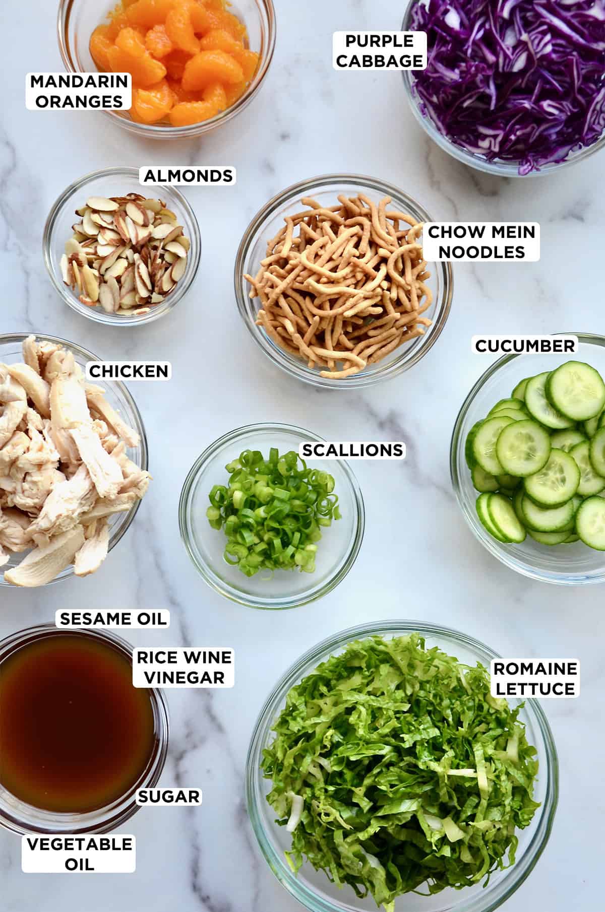Chinese chicken salad ingredients in various sizes of glass bowls, including mandarin oranges, shredded purple cabbage, crispy chow mein noodles, sliced cucumbers, chopped scallions, shredded romaine lettuce, sesame dressing, cooked chicken breast slices and sliced almonds.