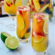 Various cocktail glasses filled with fresh fruit and white sangria in front of a pitcher filled with white sangria and a bottle of white wine.