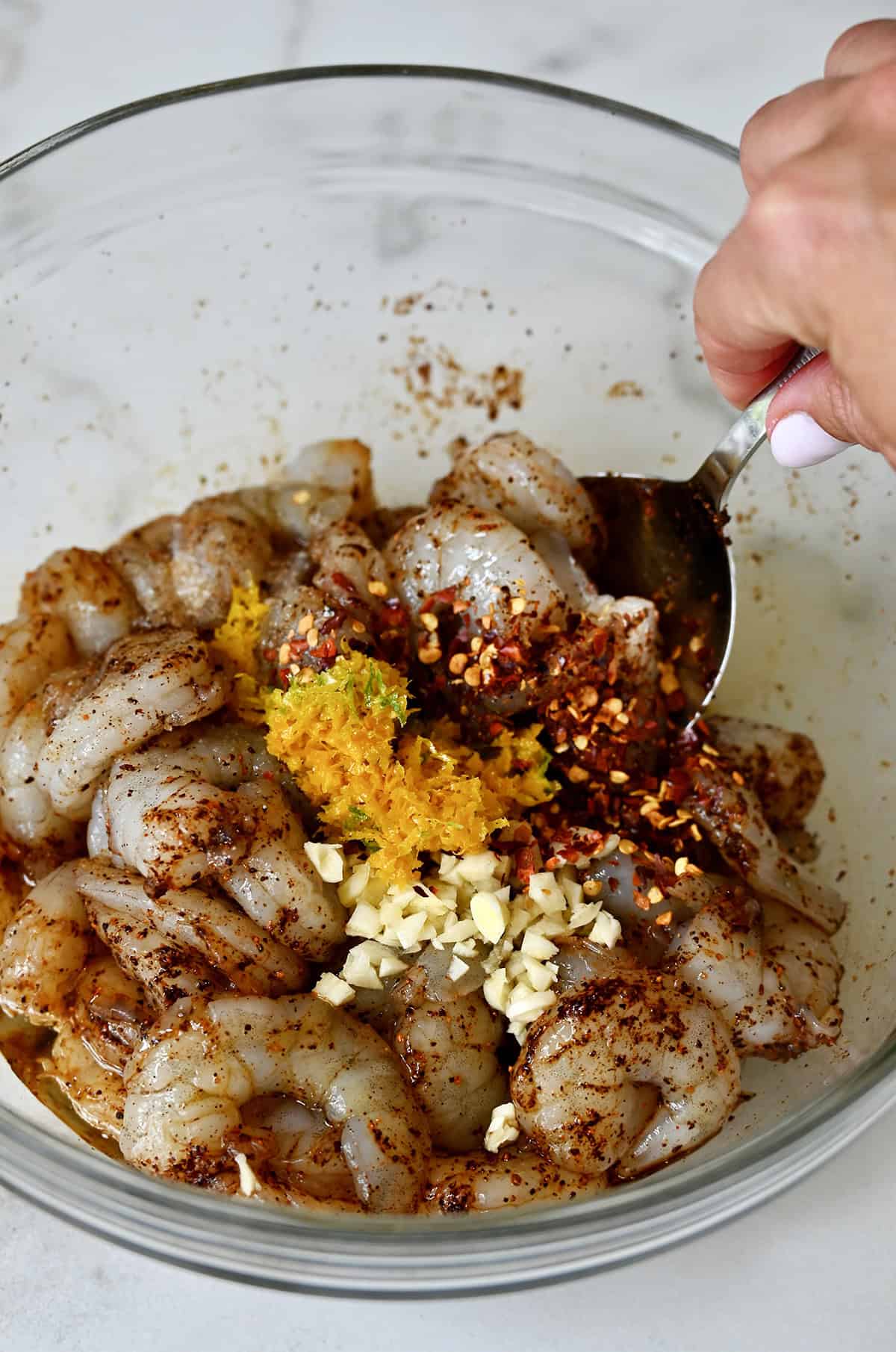 Adding orange zest, minced garlic and chili flakes to a bowl with uncooked shrimp to marinate.