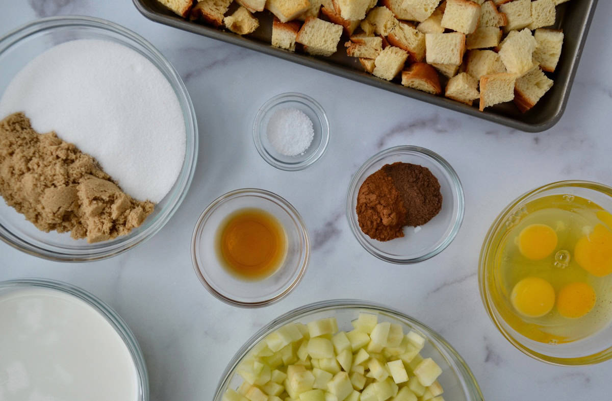 Ingredients for making apple bread pudding, including a baking sheet with cubed brioche bread, next to glass bowls containing chopped apples, pumpkin pie spice, white and brown sugar, lemon juice, half-and-half, and eggs.