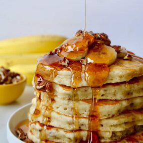 A tall stack of banana pancakes topped with bananas and maple syrup