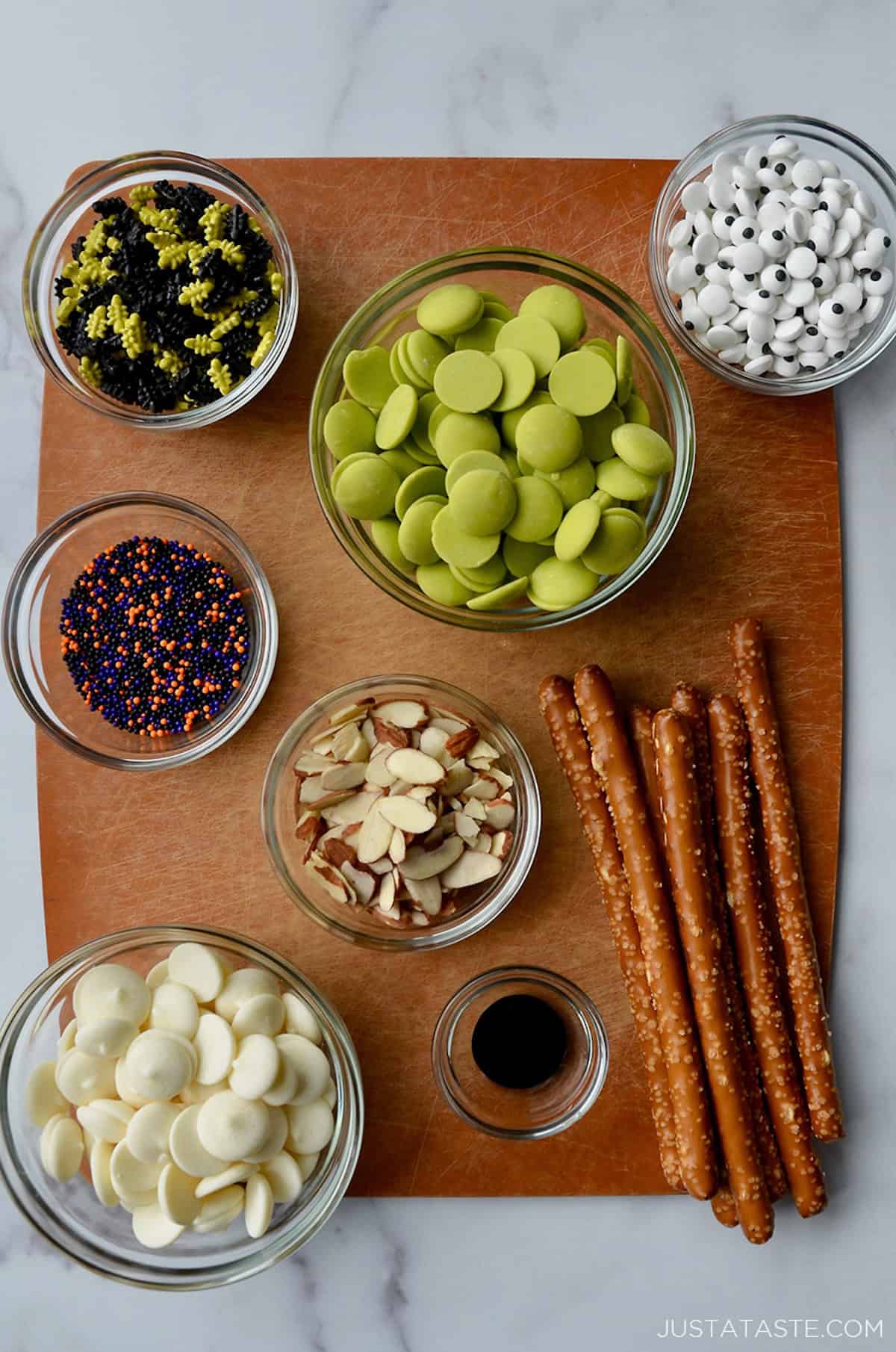 A cutting board containing glass bowls with ingredients for making chocolate-covered Halloween pretzels.