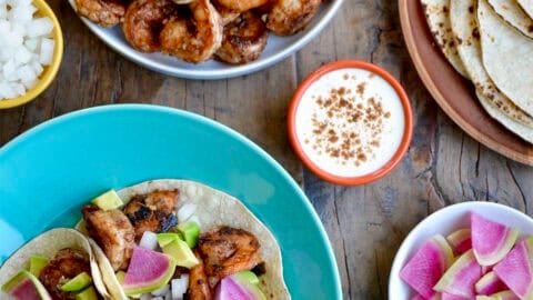 Spicy shrimp tacos on a plate next to plates piled high with spicy shrimp, charred corn tortillas, sliced radishes and diced avocado.