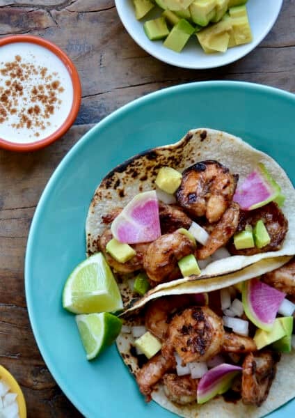 Spicy shrimp tacos in corn tortillas topped with a lime crema, sliced radishes and avocado on a dinner plate.